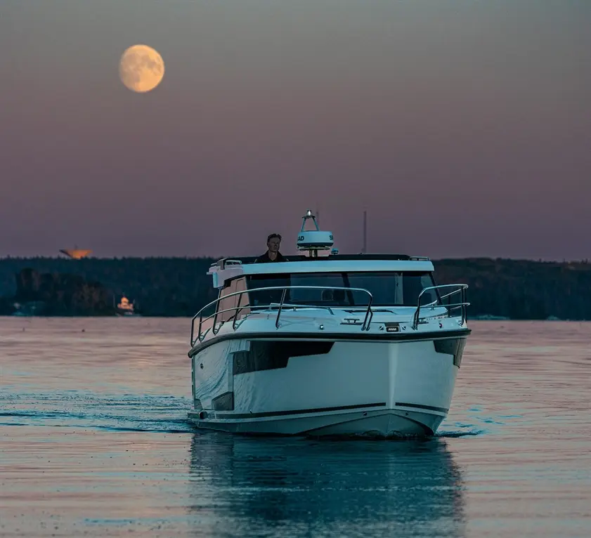 A man steers a white motorboat across a calm lake under a large moonlit sky, with silhouettes of trees in the background.